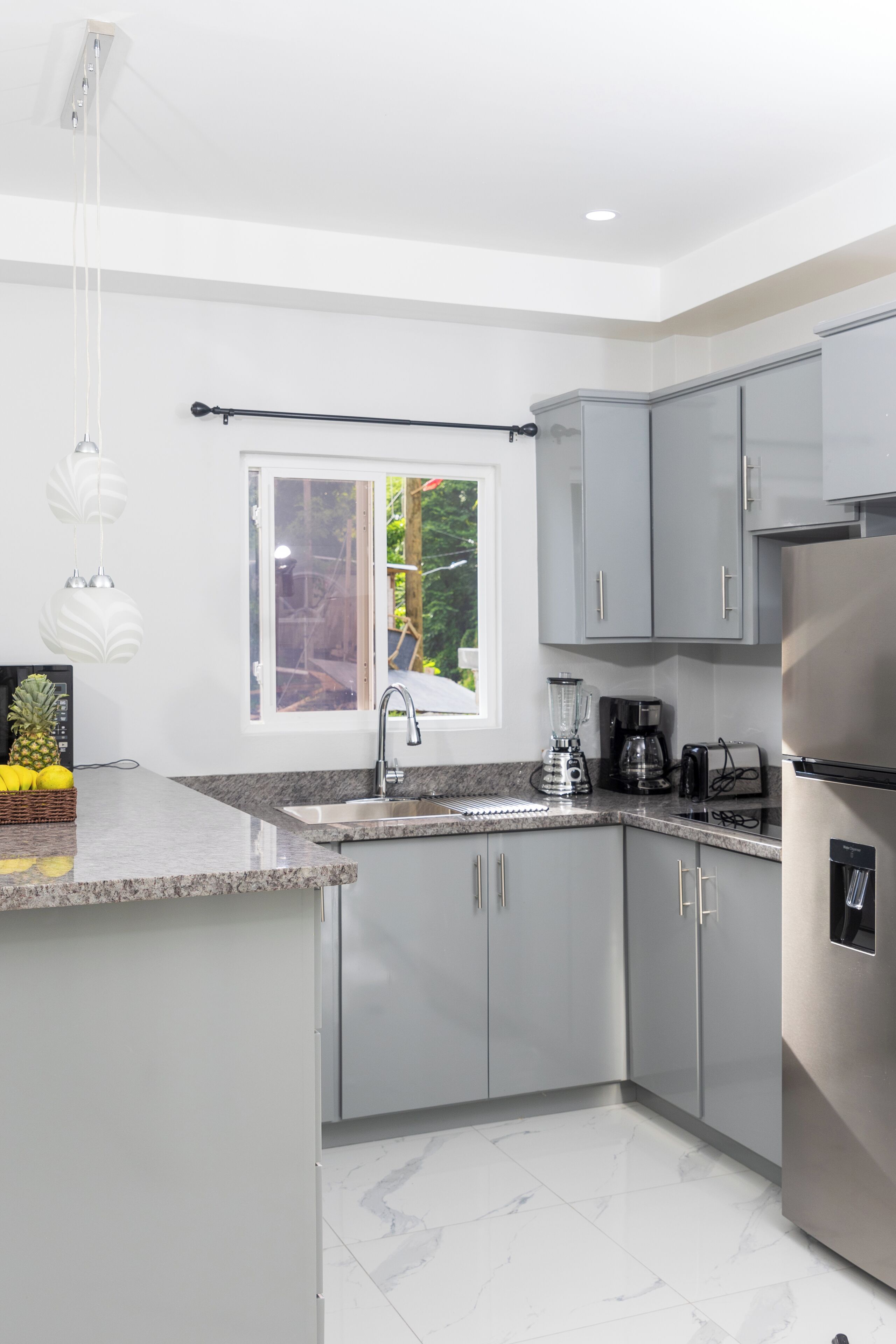 A modern kitchen with grey cabinets, a stainless steel refrigerator, a built-in oven, a sink with a faucet, and a window with a view of greenery outside.