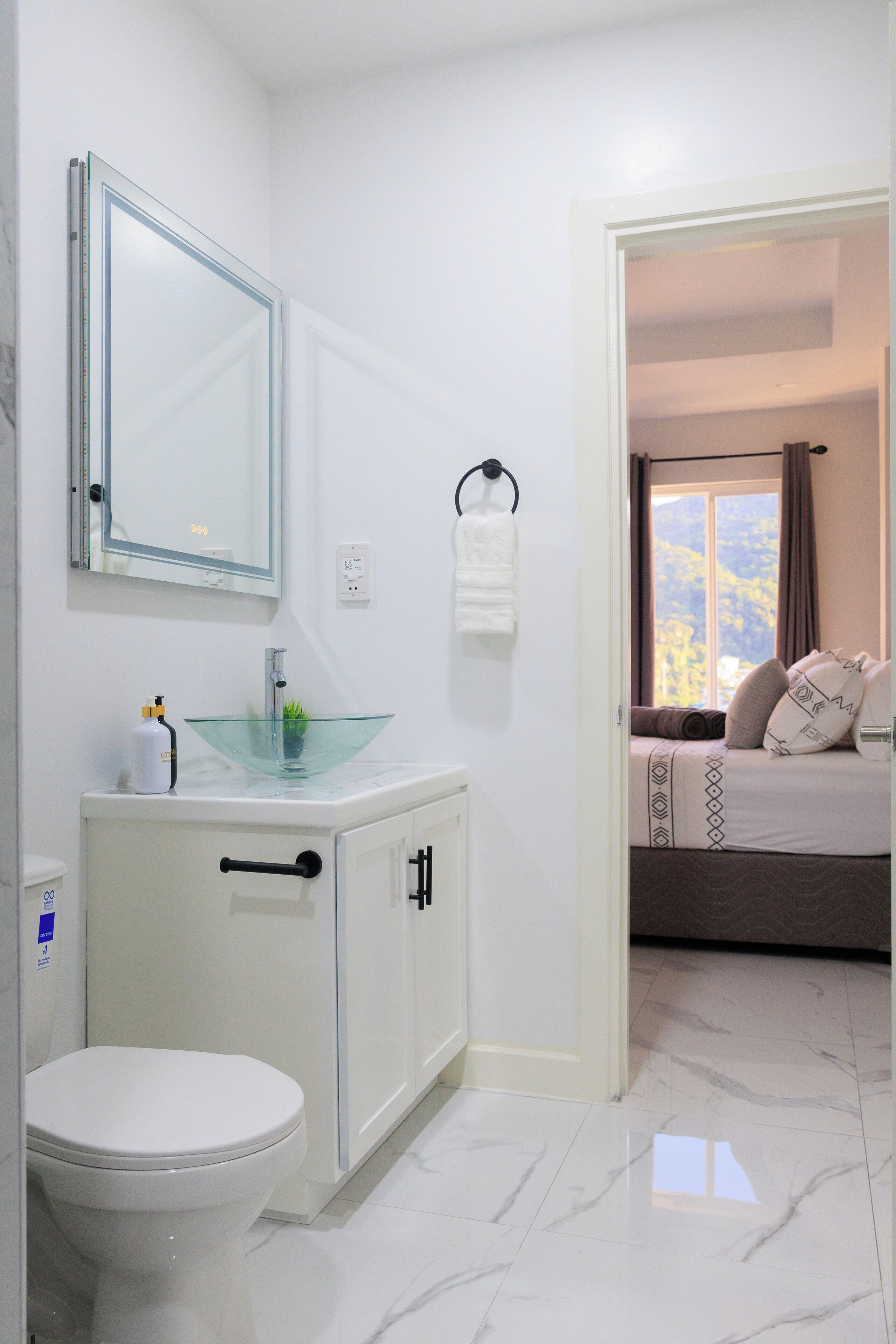 A bathroom with a white toilet, a glass sink, and a mirror. There is a towel rack and a window with curtains.