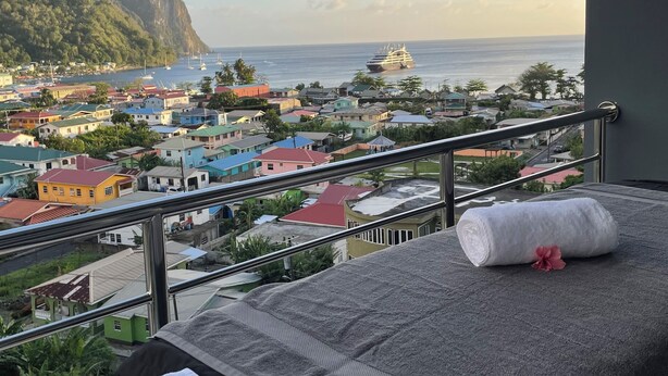 A balcony with a bed, towels, and a view of a coastal town and mountain. - Caldera Villas (Soufrière)