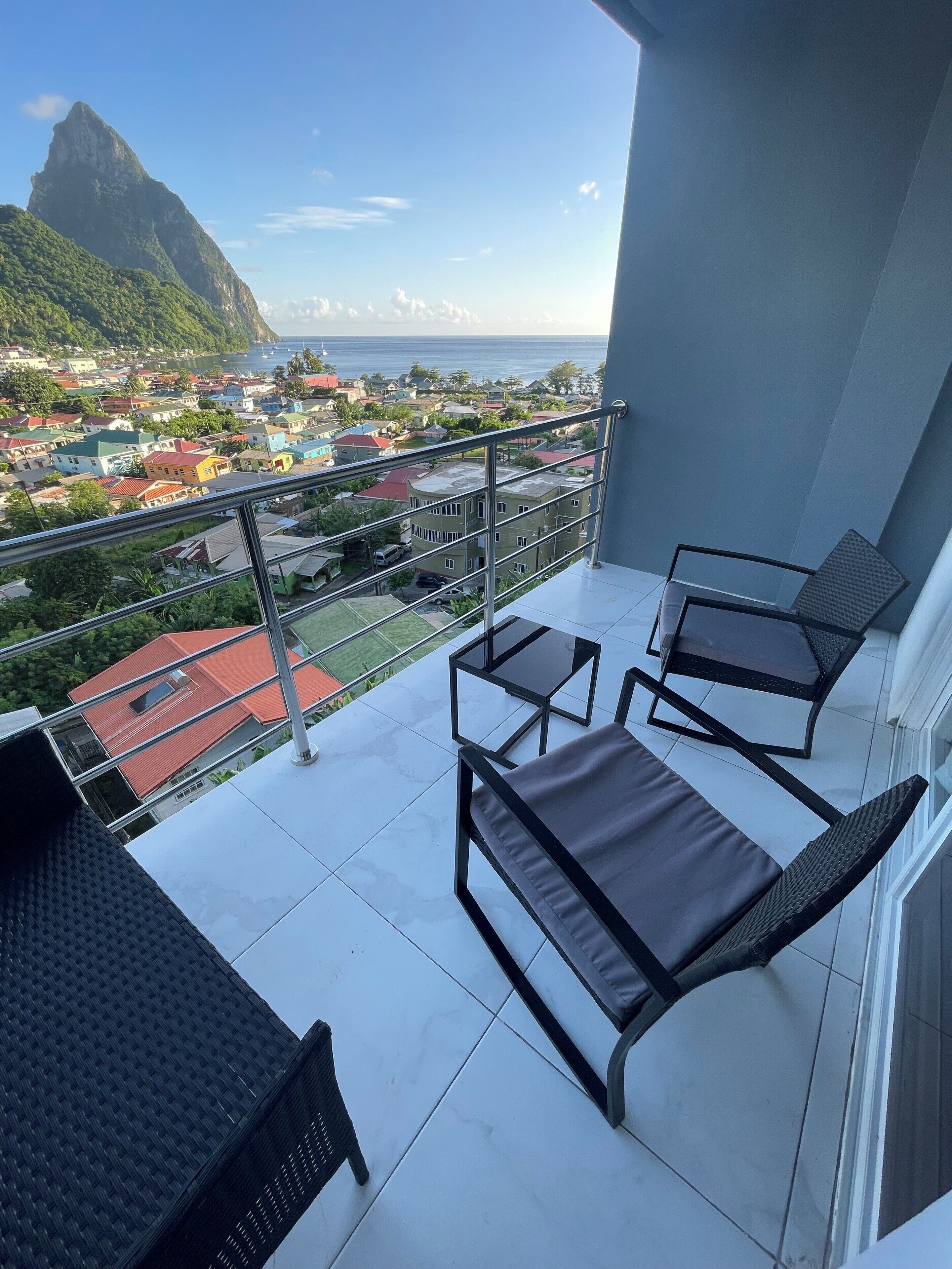 A balcony with two lounge chairs and a table, overlooking a coastal town with colorful buildings and a prominent mountain in the distance.