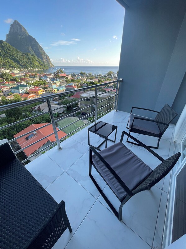 A balcony with two lounge chairs and a table, overlooking a coastal town with colorful buildings and a prominent mountain in the distance.