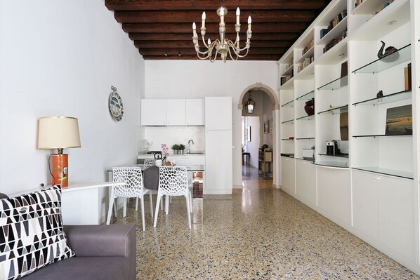 A modern living room with a sofa, a dining table, and chairs, a kitchen area with white cabinets, and a hallway leading to another room.