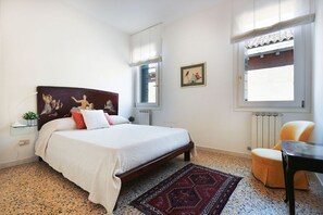A bedroom with a bed, a chair, a small table, a radiator, and a window with a view.