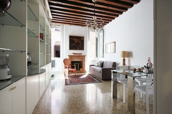 A modern kitchen with a built-in stove, a dining area with a glass table and chairs, and a living room with a sofa and fireplace.