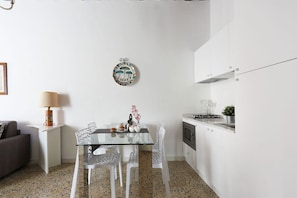 A compact kitchen with white cabinets, a glass dining table, and a decorative wall hanging.