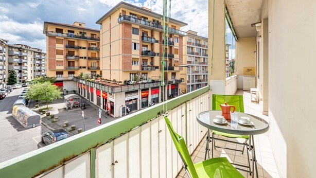 A balcony with a table and chairs, overlooking a multi-story building with balconies and a street below. - GuestHost - Cozy apartment of about 60 sqm, for up to 4 people, located on the second floor of a building with a lift (some steps to access). The apartment is located in a quiet residential area, less than 10 minutes on foot from the Cascine Park, and we (Firenze)
