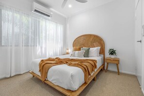 A bedroom with a bed, a bedside table, a wall-mounted air conditioner, and sheer curtains.