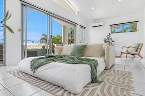 A modern bedroom with a large bed, a sofa, a wooden chair, and a view of palm trees through large windows.