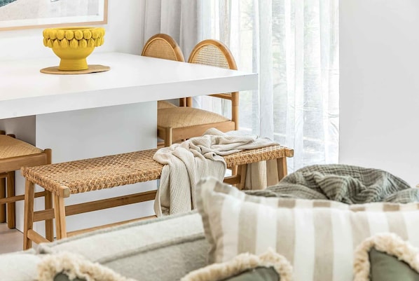 A room with a white dining table, wicker chairs, a woven bench, and a bed with striped pillows.