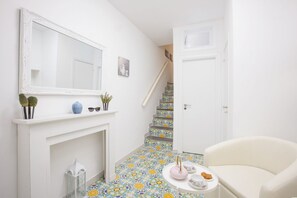 Un pasillo con una chimenea blanca, un espejo, una escalera con azulejos coloridos, una puerta blanca y un sillón blanco.