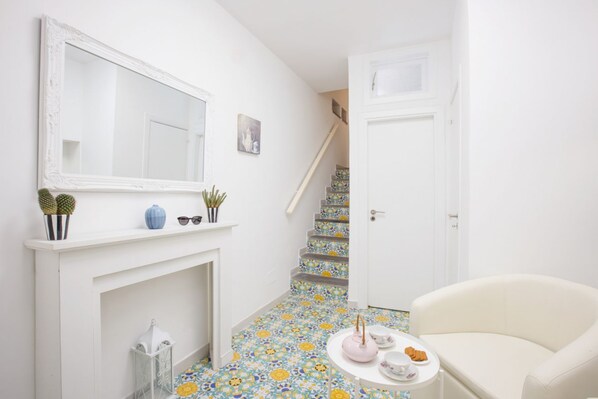 Un pasillo con una chimenea blanca, un espejo, una escalera con azulejos coloridos, una puerta blanca y un sillón blanco.