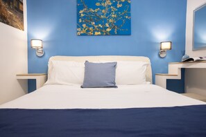Una cama bien hecha con ropa de cama blanca y azul, dos mesitas de noche con lámparas, y una pared azul con un cuadro floral.