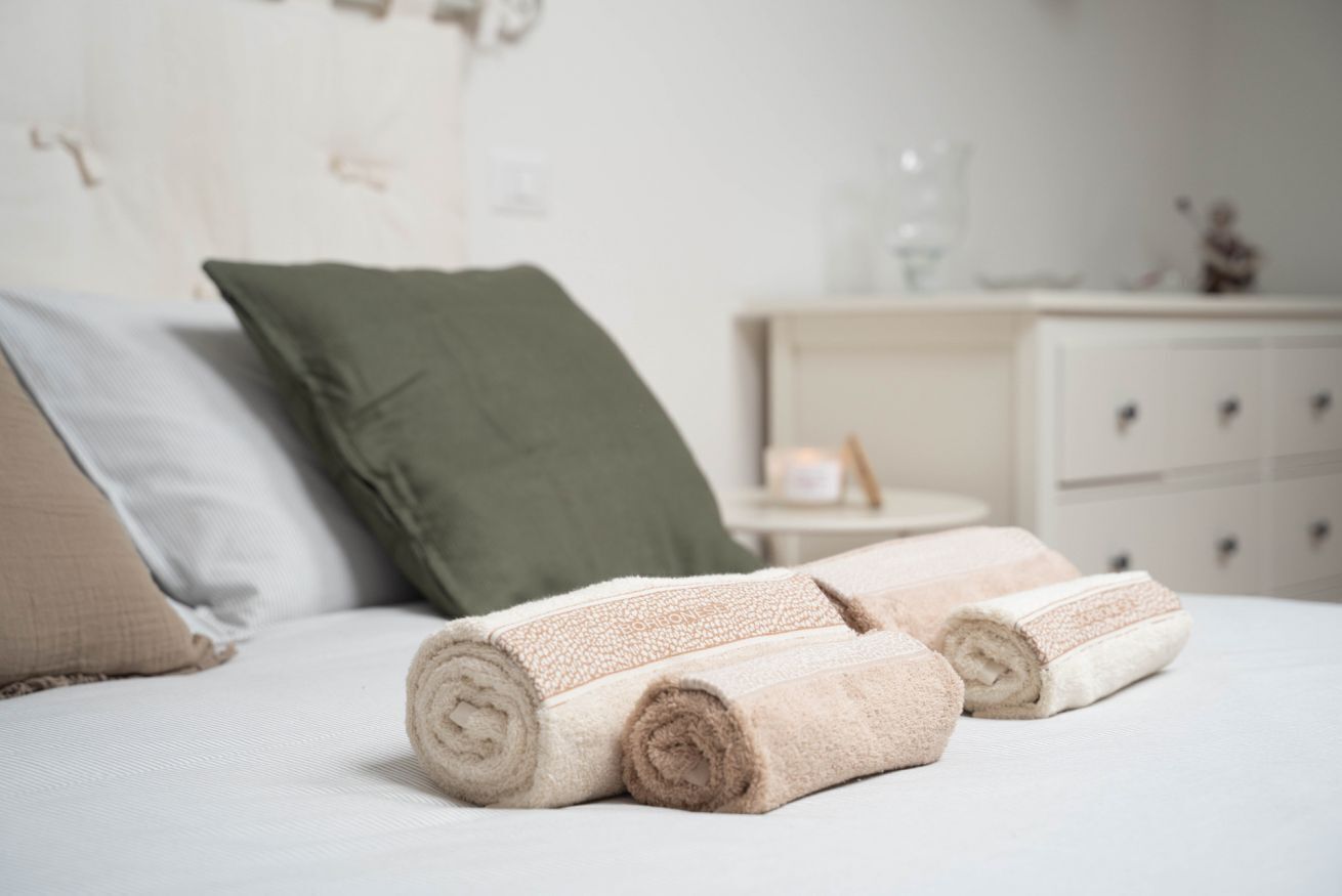 Ein ordentlich bezogenes Bett mit aufgerollten Handtüchern, einem grünen Kissen und einem Nachttisch mit Lampe und einem kleinen dekorativen Gegenstand.
