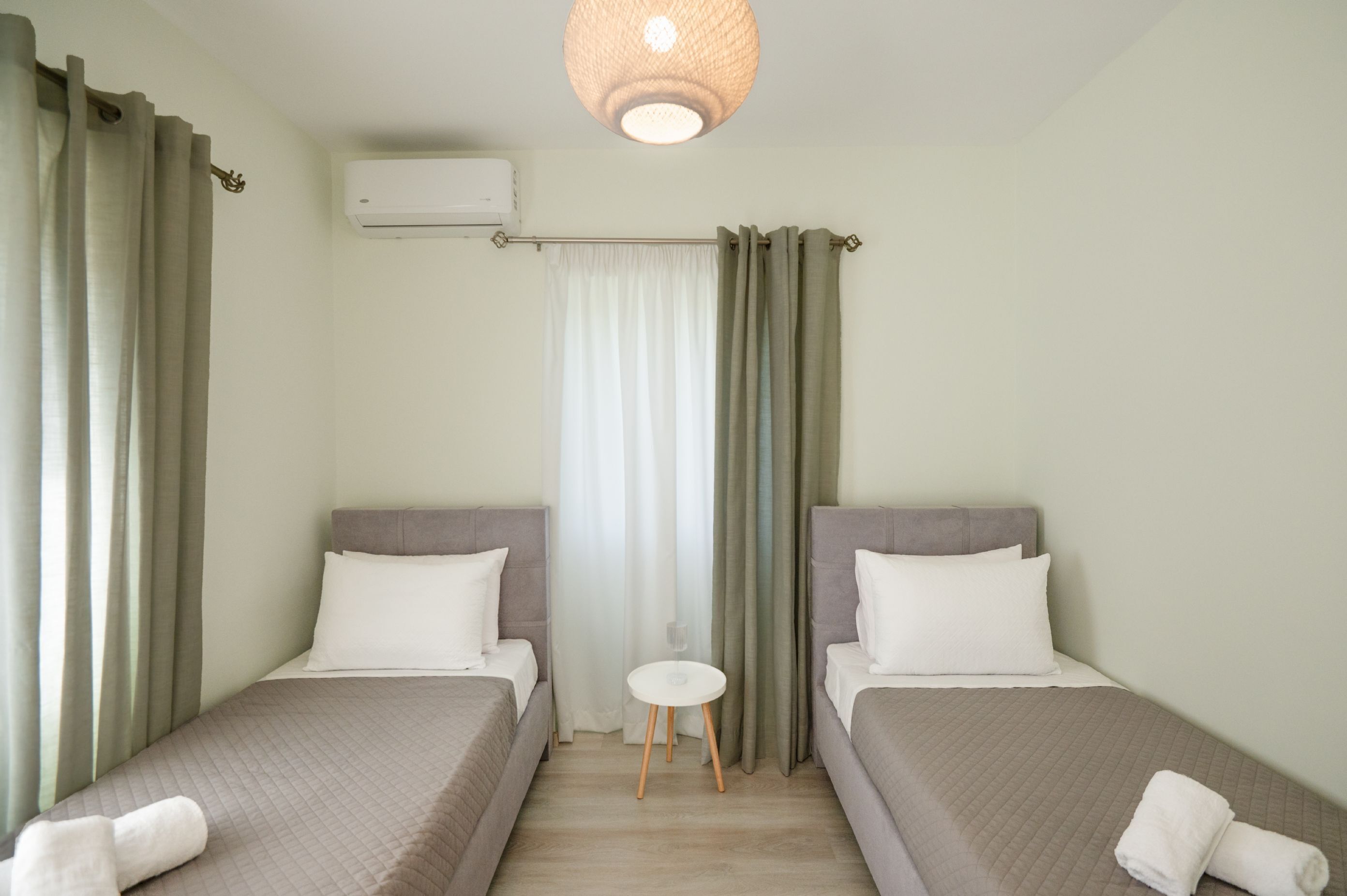 A hotel room with two single beds, a small round table, a window with curtains, and an air conditioning unit.