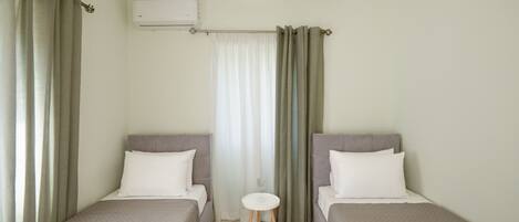 A hotel room with two single beds, a small round table, a window with curtains, and an air conditioning unit.