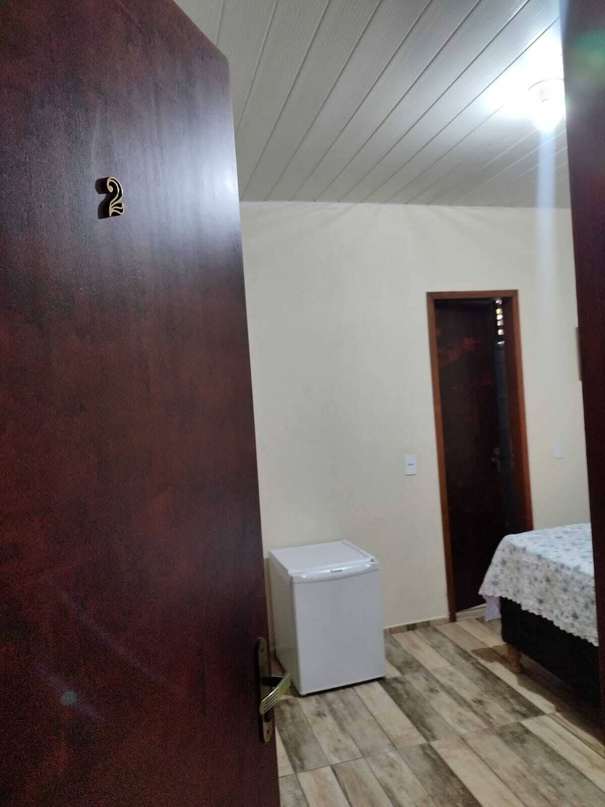 A small room with a wooden door, a white refrigerator, a bed with a floral bedspread, and a wooden floor.