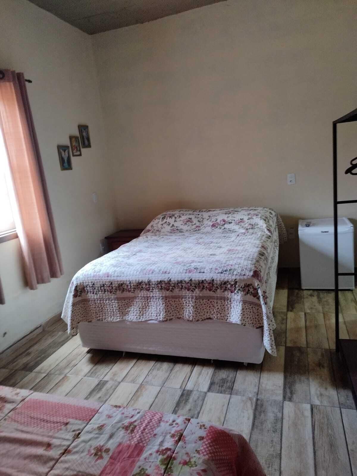 Quarto pequeno com uma cama de solteiro, colcha florida, piso de madeira, janela com cortinas e um eletrodoméstico branco pequeno à direita.