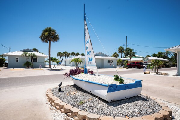 A sailboat with a sign reading "Gulf to Bay Club" in a landscaped area with palm trees and houses in the background.