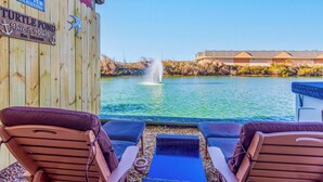 A wooden deck with two lounge chairs facing a body of water, a fountain, and a building with a lock cover in the background. - 5064 McBride's Shore Thing * 2 Min Walk to Beach (Kill Devil Hills)