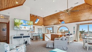 A spacious living area with a wooden ceiling, a television, a kitchen area, and a view of the outside. - 5064 McBride's Shore Thing * 2 Min Walk to Beach (Kill Devil Hills)