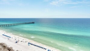 A beach with a long pier extending into the ocean, a sandy shore with umbrellas, and clear blue water. - Penthouse Paradise at The Pearl! (Navarre Beach)