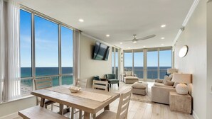 A spacious living area with a large window overlooking the ocean, a dining table, and a ceiling fan. - Penthouse Paradise at The Pearl! (Navarre Beach)