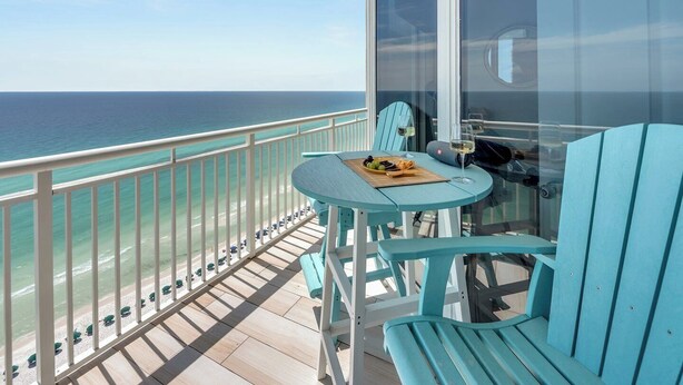 A balcony with a table set for two, overlooking a beach with umbrellas and a clear view of the ocean. - Penthouse Paradise at The Pearl! (Navarre Beach)