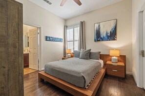 Une chambre à coucher avec un lit en bois, deux tables de chevet, une fenêtre avec des rideaux, un tableau accroché au mur et une porte donnant sur une autre pièce.