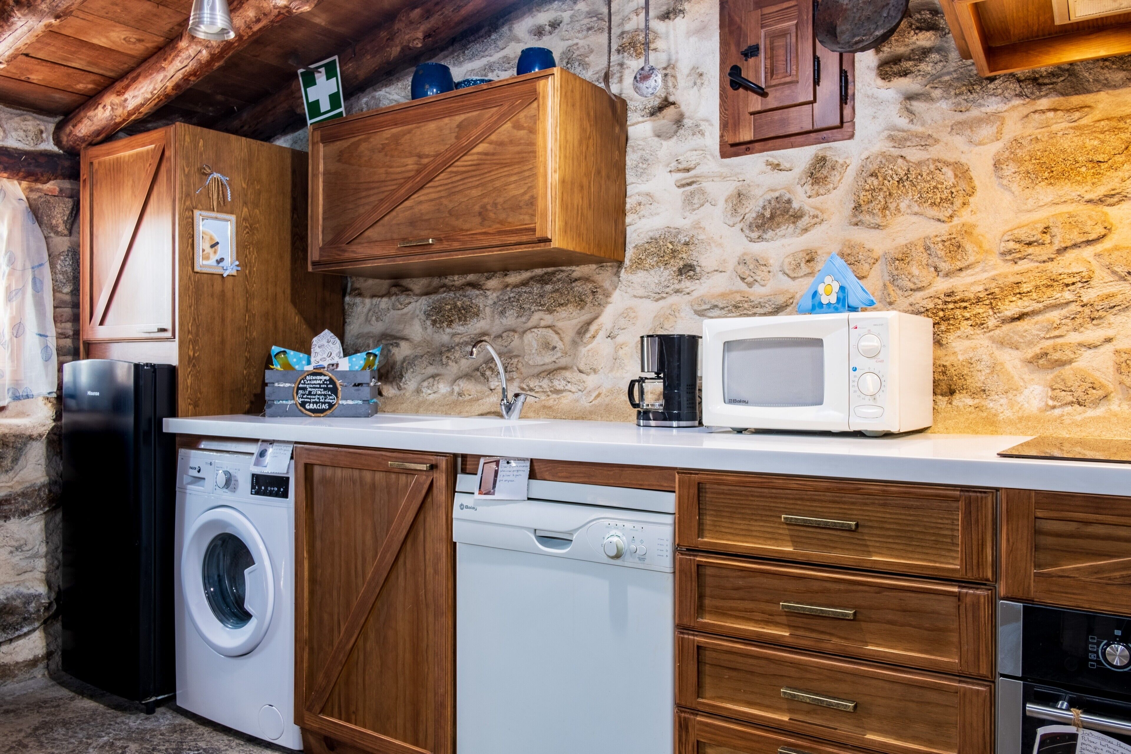 A kitchen with wooden cabinets, a washing machine, a microwave, and a stone wall.