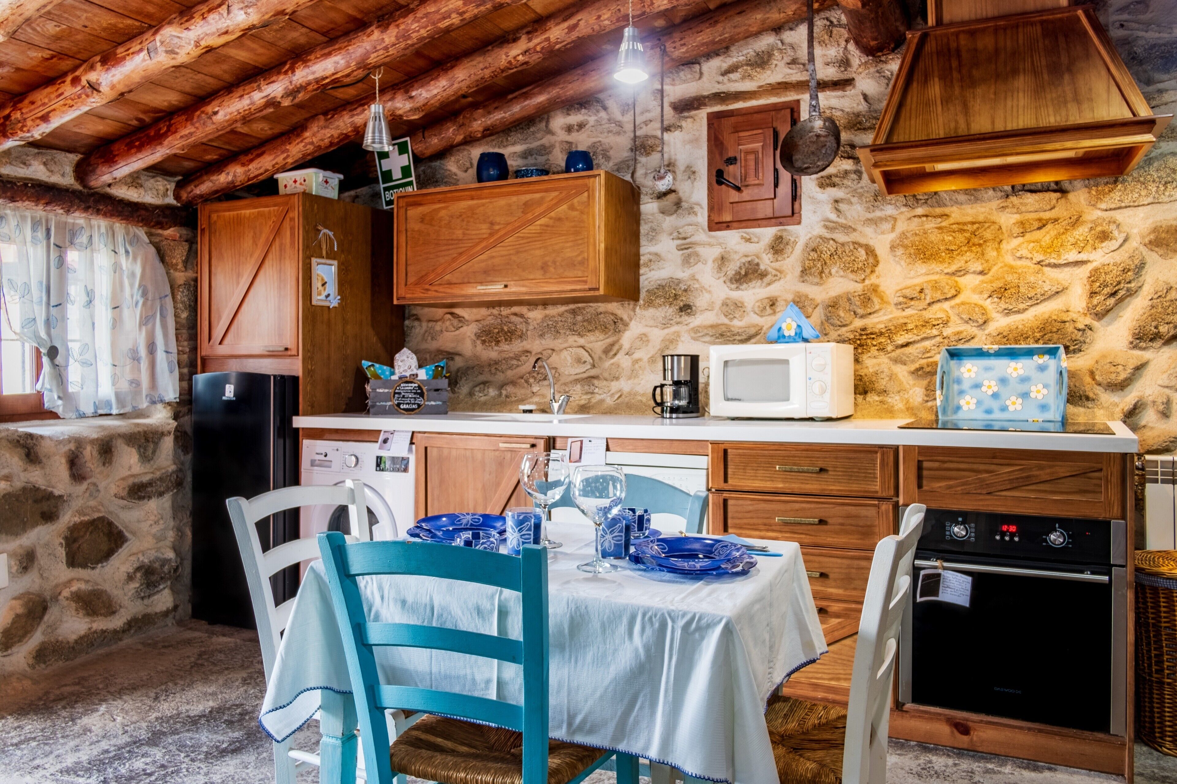 Una cocina rústica con paredes de piedra, armarios de madera, una encimera blanca y un comedor con una mesa dispuesta para dos.