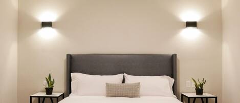 Una cama bien tendida con un cabecero gris, dos mesitas de noche con plantas en macetas, y luces fijadas a la pared.