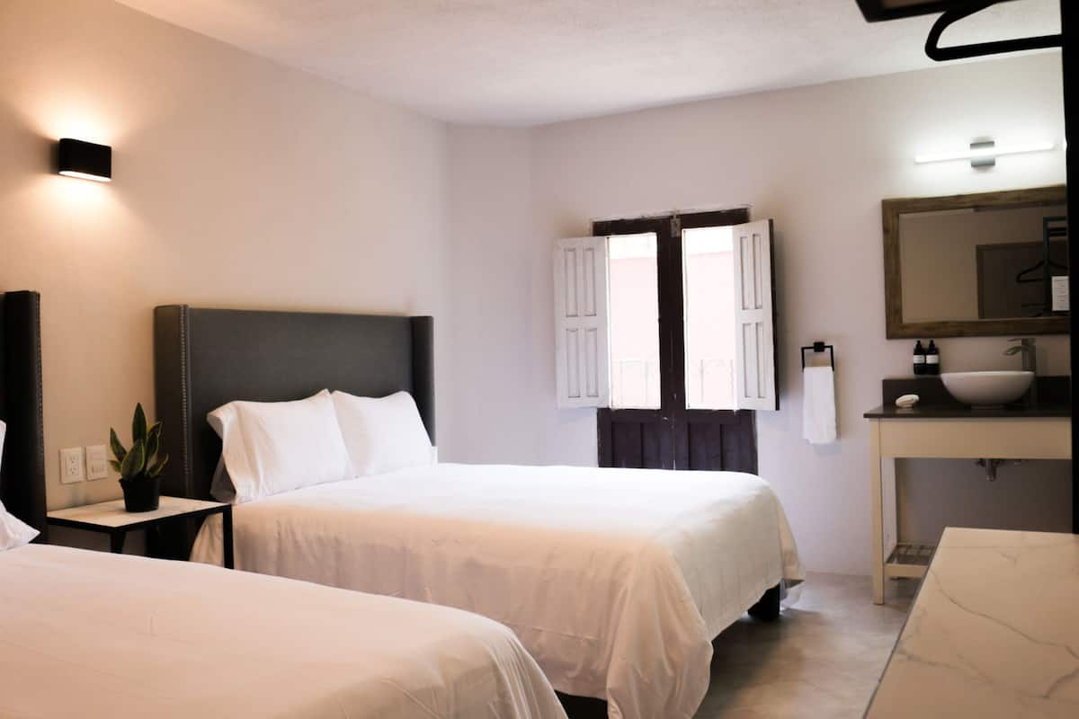Une chambre d’hôtel avec deux lits, une table de chevet agrémentée d’une plante, un grand miroir et une salle de bain avec un lavabo et une fenêtre.