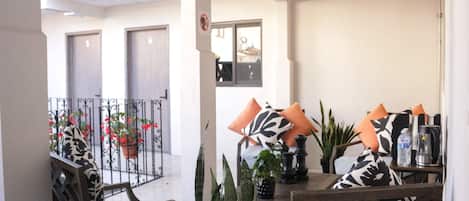 Un comedor con una mesa lista para dos personas, rodeada de sillas con cojines estampados. Hay plantas en macetas sobre la mesa y una vista a un balcón con barandilla.