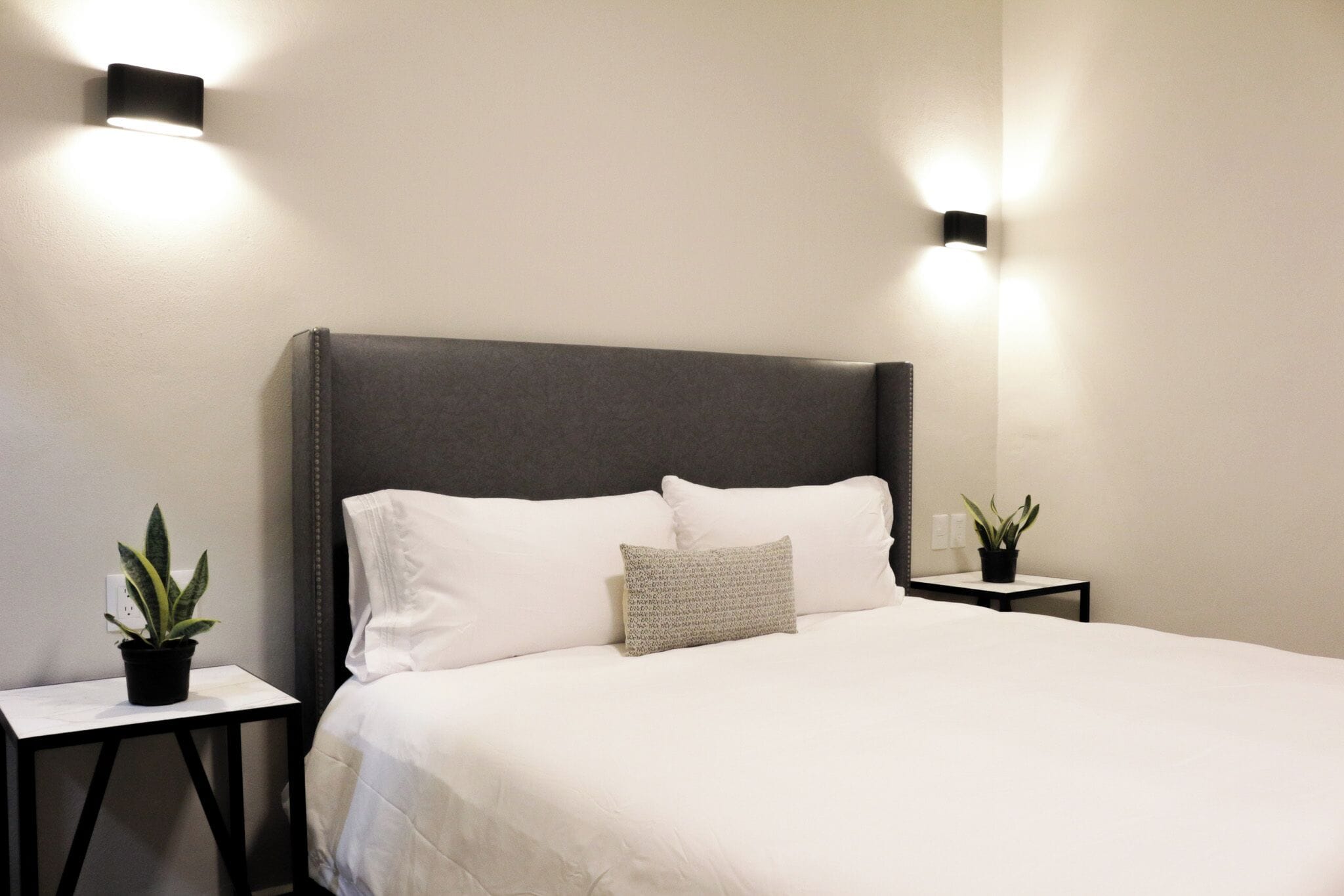 A neatly made bed with a grey headboard, two bedside tables with potted plants, and wall-mounted lights.