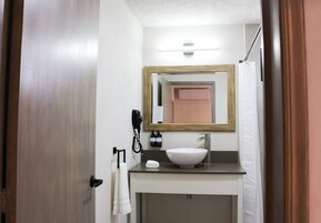 Un baño con puerta de madera, lavamanos blanco, espejo y toallero.
