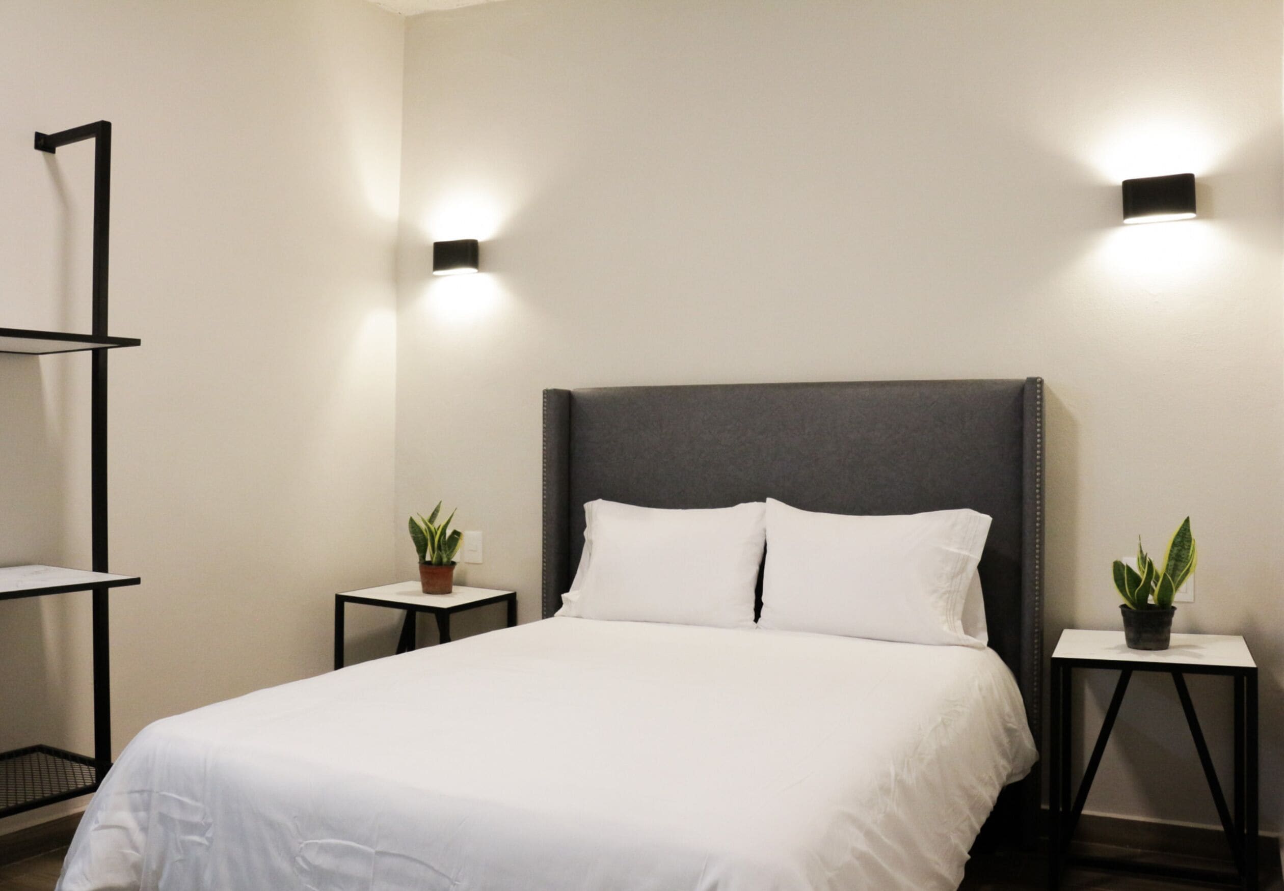 A neatly made bed with white linens, two bedside tables with potted plants, and wall-mounted lighting.