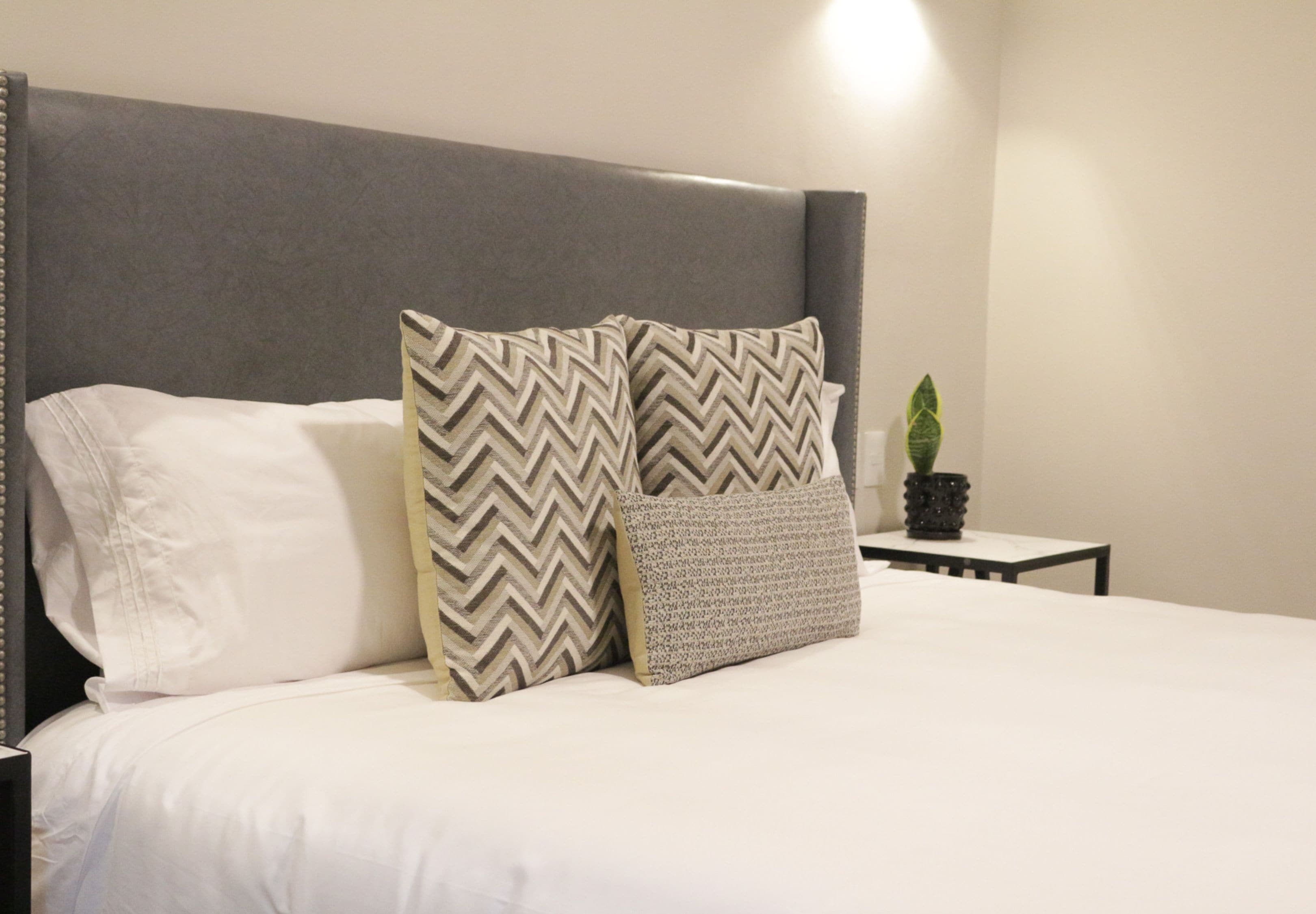 A neatly made bed with a grey upholstered headboard, two patterned pillows, and a small potted plant on a bedside table.