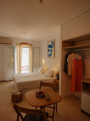 A hotel room with a bed, a dining table with a chair, a small table with a cup and a plate of fruit, a wardrobe, and a window with curtains.