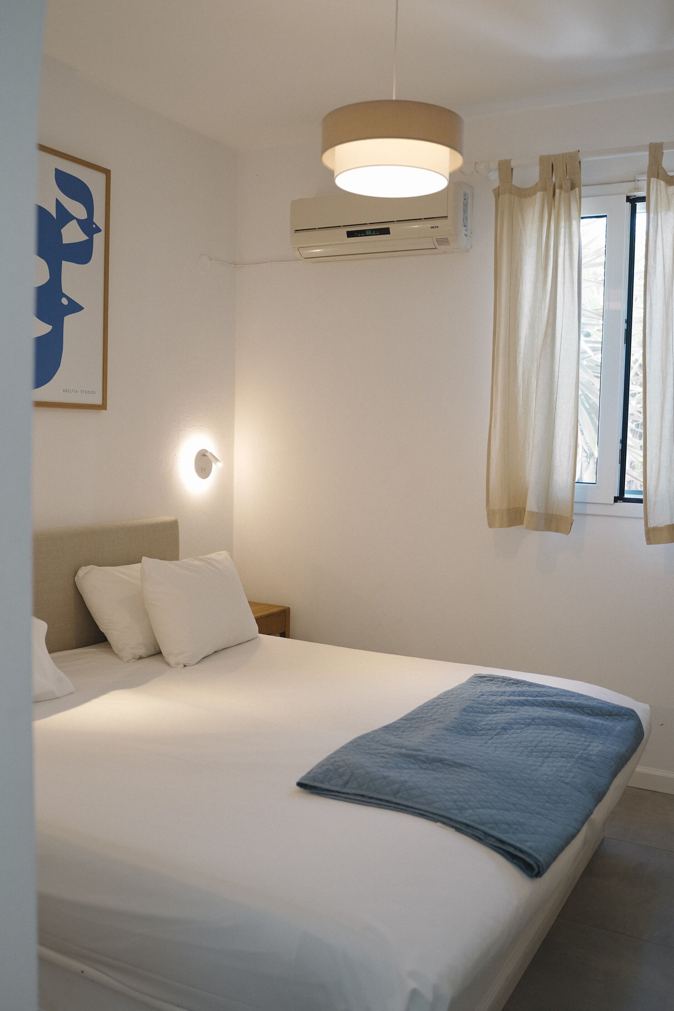 Una cama bien hecha con sábanas blancas y una manta azul, un aire acondicionado de pared, una lámpara de noche y una ventana con cortinas.