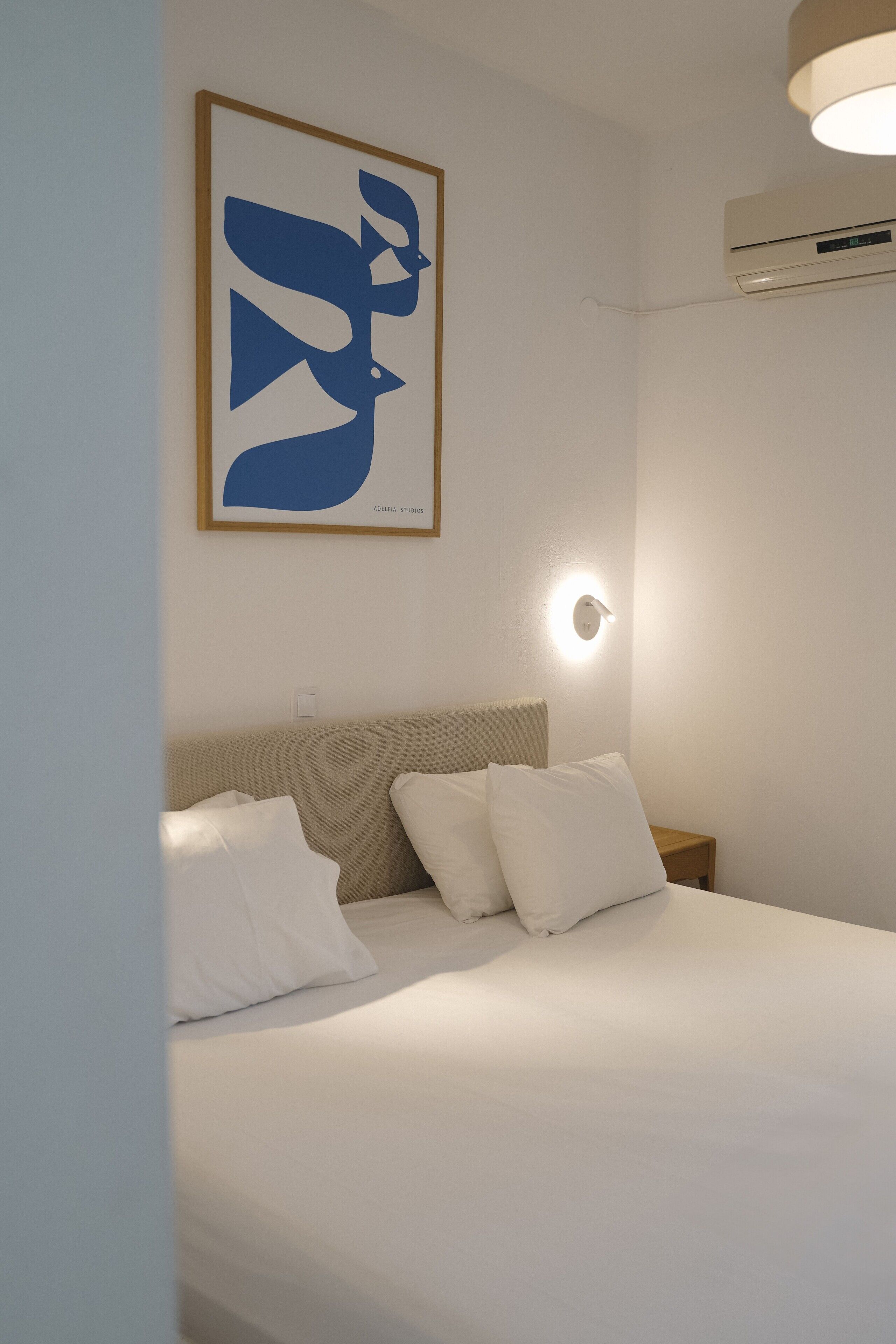 Une chambre à coucher avec un lit, des oreillers blancs, une œuvre d’art accrochée au mur, une table de chevet et un luminaire fixé au mur.