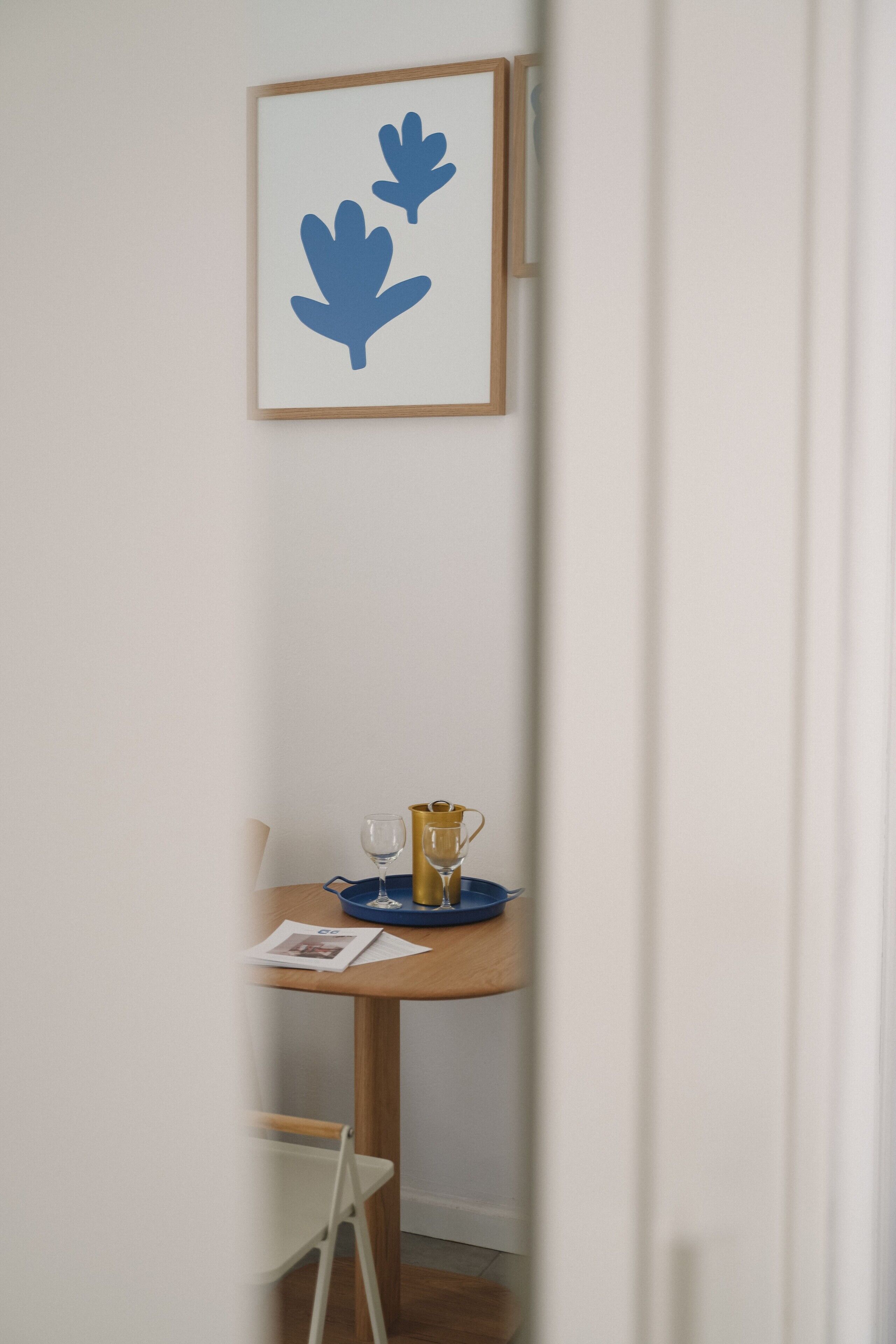 Un pasillo estrecho con una obra enmarcada en la pared, una mesita con una copa, una taza y una revista, y una silla blanca.