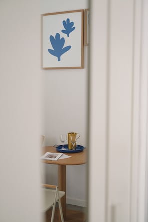 A narrow hallway with a framed artwork on the wall, a small table with a glass, a cup, and a magazine, and a white chair.