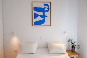 A bedroom with a bed, two pillows, a nightstand with a plant, and a framed abstract artwork on the wall.