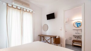Une chambre d’hôtel comprenant un lit, un bureau, une chaise, une télévision, un miroir rond, une salle de bain avec un lavabo et des toilettes, ainsi qu’un espace douche.