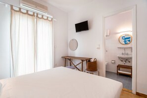 A hotel room with a bed, a desk, a chair, a TV, a round mirror, a bathroom with a sink and toilet, and a shower area.