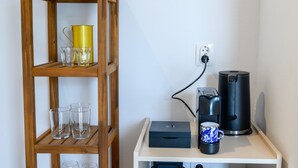 Une étagère en bois avec des verres et un récipient jaune, une table blanche avec une machine à café et une tasse, et une prise électrique sur le mur.