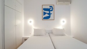 Une chambre avec deux lits, du linge de lit blanc, un climatiseur fixé au mur et une œuvre d’art abstraite encadrée.