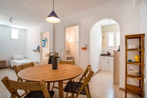 A dining area with a round wooden table, chairs, a hanging light fixture, and a kitchen visible through an archway.