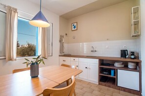 A compact kitchen with a dining table, chairs, a hanging light fixture, a window with curtains, and a view of buildings outside.