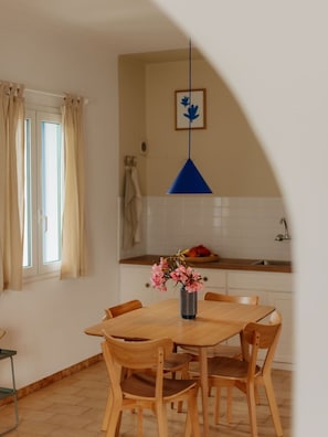 A dining area with a wooden table and chairs, a vase with flowers, a blue pendant light, and a window with curtains.